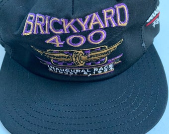 Könnte beinhalten: Schwarze Baseballkappe mit einem gestickten Logo in Lila und Gold, auf dem "Brickyard 400" und "Inaugural Race August 21, 1994" steht.