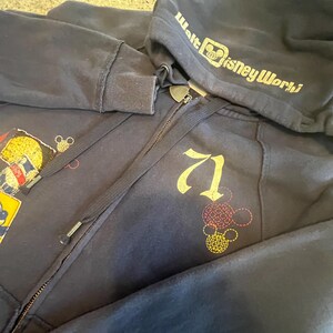 May include: A navy blue zip-up hoodie featuring the text Walt Disney World on the hood and the number 71 printed on the chest. The garment includes embroidered Mickey Mouse head outlines in red and yellow, along with a patch on the left side.