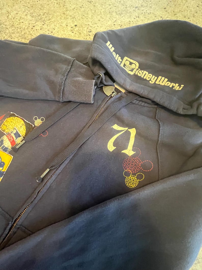 May include: A navy blue zip-up hoodie featuring the text Walt Disney World on the hood and the number 71 printed on the chest. The garment includes embroidered Mickey Mouse head outlines in red and yellow, along with a patch on the left side.