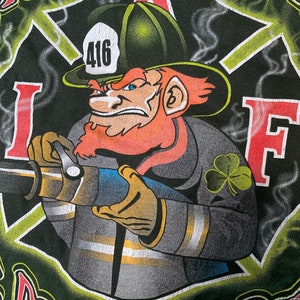 Puede incluir: Una camiseta gráfica verde, negra y amarilla con un duende bombero de dibujos animados sosteniendo una manguera. El duende lleva un sombrero verde con una banda roja y un uniforme de bombero negro y amarillo. La camiseta tiene el texto "Irish Ol' 416" y "Patrick's Day".
