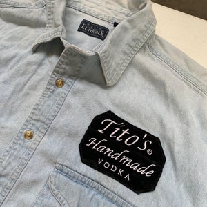 May include: Light blue denim shirt with a black patch that says "Tito's Handmade Vodka" on it. The shirt has a Basic Editions label on the collar.