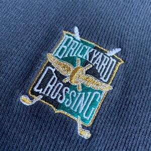 May include: A navy blue embroidered patch with a green and gold shield. The shield has the text "Brickyard Crossing" on it. The shield is surrounded by two crossed golf clubs.