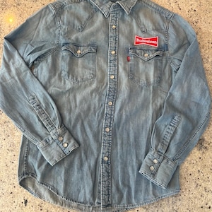 Może przedstawiać: Jasnoniebieska koszula dżinsowa z długimi rękawami, dwiema kieszeniami na piersi i zapięciem na guziki. Posiada czerwoną naszywkę Budweiser na prawej piersi i małą czerwoną metkę Levi's. Klasyczny styl western.