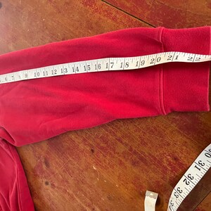 May include: Red long-sleeve shirt with a folded cuff. A measuring tape is laid across the sleeve, showing a measurement of 21 inches.
