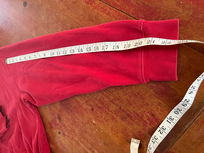 May include: Red long-sleeve shirt with a folded cuff. A measuring tape is laid across the sleeve, showing a measurement of 21 inches.