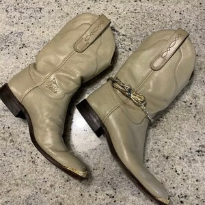May include: A pair of light grey leather cowboy boots with gold accents. The boots have a pointed toe and a decorative stitching detail on the shaft.