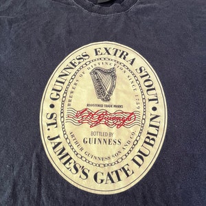 Puede incluir: Camiseta negra con un logotipo vintage de Guinness Extra Stout. El logotipo es un c&iacute;rculo dorado con un arpa, las palabras "Brewers of Distinction Since 1759", y "St. James's Gate Dublin".