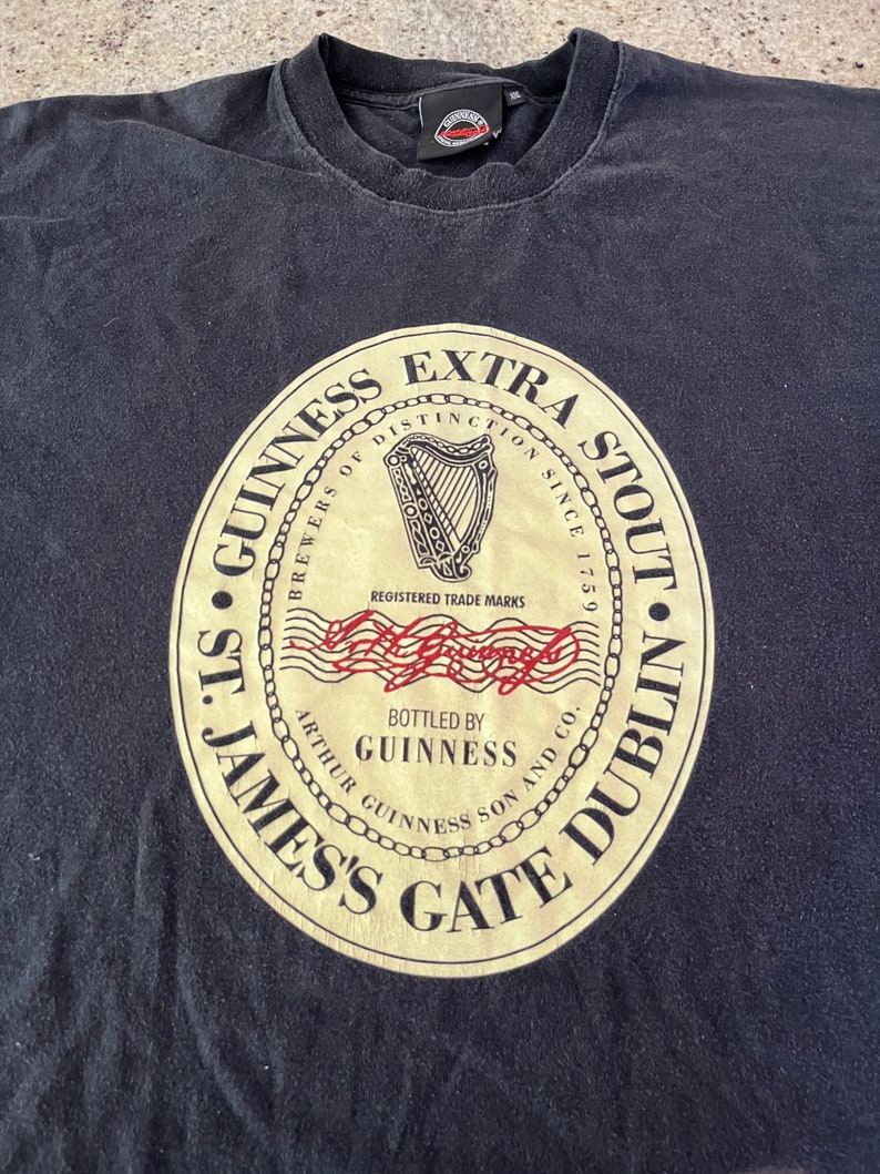 Puede incluir: Camiseta negra con un logotipo vintage de Guinness Extra Stout. El logotipo es un c&iacute;rculo dorado con un arpa, las palabras "Brewers of Distinction Since 1759", y "St. James's Gate Dublin".