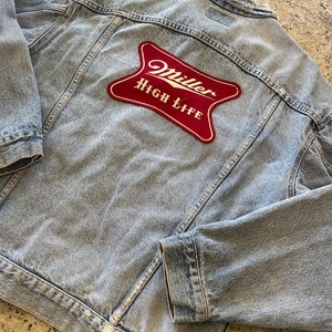 Puede incluir: Una chaqueta vaquera azul claro con un parche rojo de Miller High Life en la espalda.
