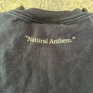 Puede incluir: Camiseta negra con el texto "Natural Anthem." impreso en amarillo en la parte posterior.
