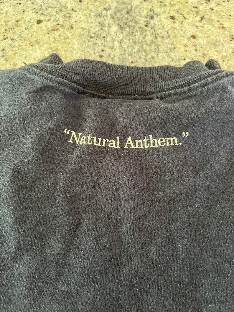 Puede incluir: Camiseta negra con el texto "Natural Anthem." impreso en amarillo en la parte posterior.
