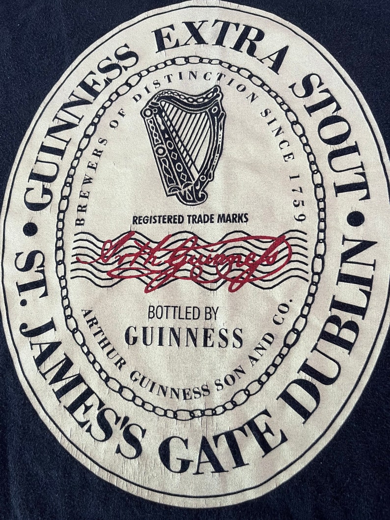 Puede incluir: Una etiqueta de cerveza Guinness Extra Stout de estilo vintage con un logotipo de arpa en blanco y negro, el texto "Brewers of Distinction Since 1759" y las palabras "St. James's Gate Dublin" en un dise&ntilde;o circular.