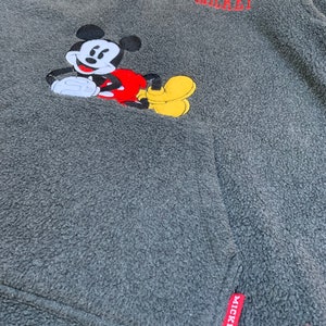 May include: Gray fleece pullover with a red and yellow embroidered Mickey Mouse design. The zipper has a gold pull and the bottom of the zipper has a red tag with "MICKEY" written on it. The top of the pullover has "MICKEY" embroidered in red.