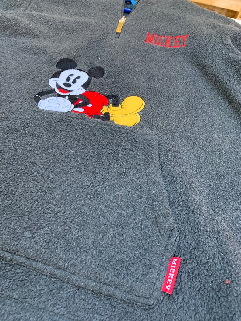 May include: Gray fleece pullover with a red and yellow embroidered Mickey Mouse design. The zipper has a gold pull and the bottom of the zipper has a red tag with "MICKEY" written on it. The top of the pullover has "MICKEY" embroidered in red.