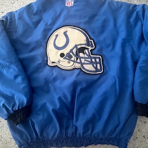Puede incluir: Una chaqueta azul con ribetes negros en el cuello y los puños. La parte trasera presenta un gran logotipo bordado del casco de los Indianapolis Colts en blanco y azul, con un pequeño parche del logo de la NFL sobre el casco.