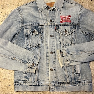 Puede incluir: Una chaqueta vaquera azul claro con dobladillo desgastado y un parche rojo y blanco de Miller High Life en el bolsillo izquierdo.