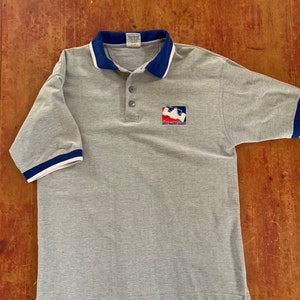 May include: Grey short-sleeved polo shirt with a blue and white collar and cuffs. The shirt has a logo on the front that says "Indy Racing League".