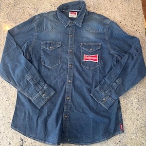 May include: A long-sleeved denim shirt with a button-down front, two chest pockets, and a classic collar. The shirt features the Budweiser logo on the right chest and a small red tag on the lower left side. The shirt is a medium blue wash.