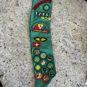 May include: A green Girl Scout sash with various badges and patches, including "Girl Scouts USA", "Hoosier Capital", "Bridge to Juniors", "Junior Aide", and a number "1225".