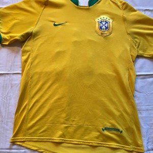 以下が含まれることがあります： 緑色のトリムとナイキのスウッシュロゴが付いた黄色のサッカーユニフォーム。ユニフォームには、「BRASIL」という文字と5つの星がついたブラジルのナショナルチームの紋章が描かれています。
