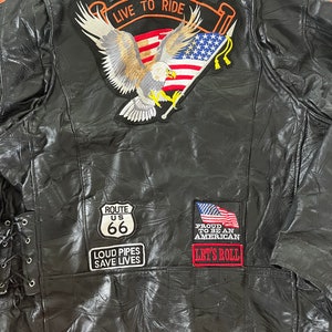Puede incluir: Chaqueta de cuero negra para motocicleta con varios parches. Los parches incluyen un águila con una bandera, un letrero de la Ruta 66, un parche de "Loud Pipes Save Lives" y un parche de "Proud to be an American, Let's Roll".