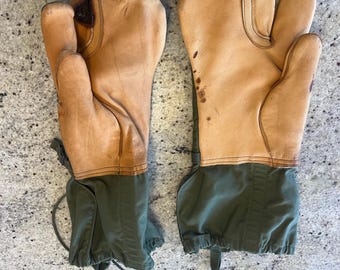 May include: A pair of brown leather mittens with green fabric cuffs and drawstrings. The mittens have a fingerless design and are worn.