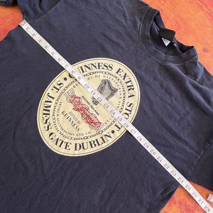 Puede incluir: Camiseta negra con un logotipo dorado de Guinness Extra Stout. El logotipo presenta un arpa, las palabras "Guinness Extra Stout" y "St. James's Gate Dublin".