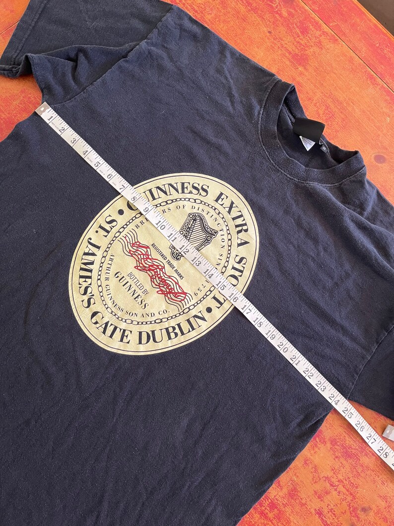 Puede incluir: Camiseta negra con un logotipo dorado de Guinness Extra Stout. El logotipo presenta un arpa, las palabras "Guinness Extra Stout" y "St. James's Gate Dublin".