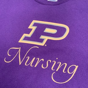 May include: A purple sweatshirt with the word "Nursing" in a cursive font below a large "P" logo with a gold and gray outline. The sweatshirt is made of a soft material and has a crew neck.