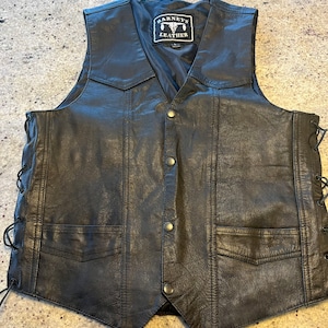 Può includere: Gilet di pelle nera con dettagli di pizzo sui lati. Il gilet ha un'etichetta "BARNEY'S LEATHER" con un logo a testa di toro.