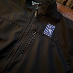 Puede incluir: Chaqueta softshell negra con cierre de cremallera y un parche con el logotipo de Rolls Royce azul en el pecho.