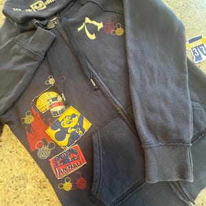 May include: A dark navy blue zip-up hoodie featuring a Walt Disney World logo on the hood. The front displays various colorful patches, including a yellow Mickey Mouse silhouette, a red Animal Kingdom logo, and several circular Mickey icon outlines.