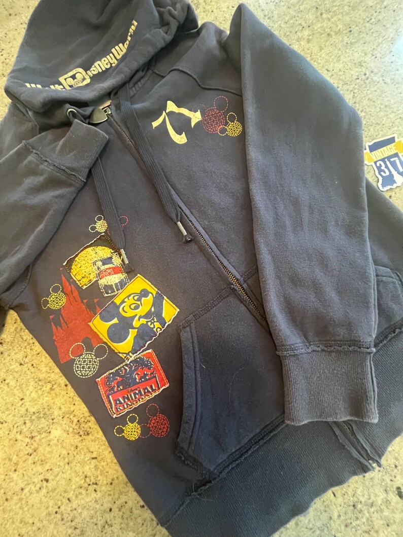 May include: A dark navy blue zip-up hoodie featuring a Walt Disney World logo on the hood. The front displays various colorful patches, including a yellow Mickey Mouse silhouette, a red Animal Kingdom logo, and several circular Mickey icon outlines.