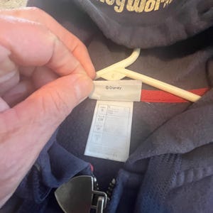 May include: A navy blue zip-up hoodie featuring the text Disney World in a distressed gold font on the hood. The garment includes a silver-toned metal zipper pull shaped like a shield and a white interior tag labeled Disney with size S.