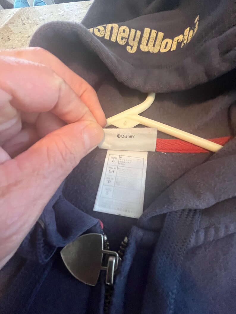 May include: A navy blue zip-up hoodie featuring the text Disney World in a distressed gold font on the hood. The garment includes a silver-toned metal zipper pull shaped like a shield and a white interior tag labeled Disney with size S.