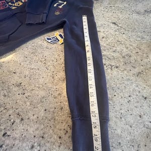 May include: A navy blue zip-up hoodie with a hood, long sleeves, and ribbed cuffs. A white measuring tape is placed along the sleeve, showing a length of approximately 21 inches. The hoodie features a graphic print on the chest and a small tag.
