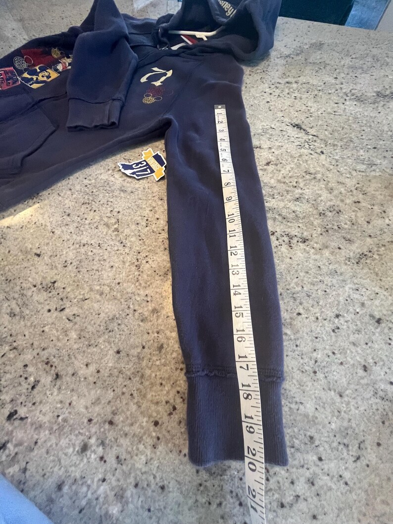 May include: A navy blue zip-up hoodie with a hood, long sleeves, and ribbed cuffs. A white measuring tape is placed along the sleeve, showing a length of approximately 21 inches. The hoodie features a graphic print on the chest and a small tag.