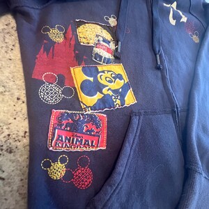 May include: A navy blue zip-up hoodie featuring a variety of patches and prints. The design includes a yellow square patch with Mickey Mouse, a rectangular patch with the text Disney Animal Kingdom, and several red and yellow Mickey Mouse head icons.