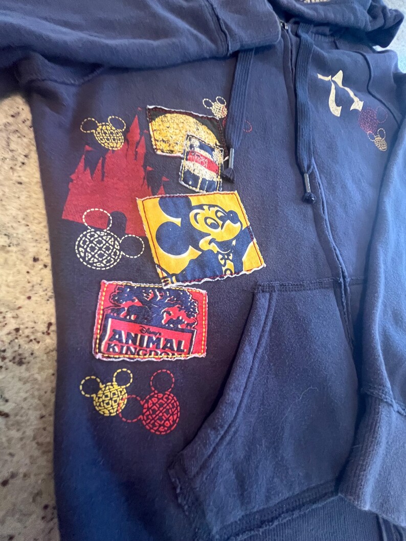 May include: A navy blue zip-up hoodie featuring a variety of patches and prints. The design includes a yellow square patch with Mickey Mouse, a rectangular patch with the text Disney Animal Kingdom, and several red and yellow Mickey Mouse head icons.