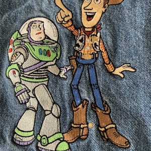 Puede incluir: Parche de chaqueta vaquera bordada con Woody y Buzz Lightyear de la franquicia Toy Story. Woody lleva un sombrero de vaquero marrón, una camisa de cuadros rojos y amarillos y pantalones vaqueros azules. Buzz Lightyear lleva un traje espacial verde y blanco con un casco morado. El parche está sobre un fondo de mezclilla azul.