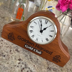 May include: A wooden clock with a white face and black hands. The clock is on a wooden base with the words "Golden Harvest Gold Club" engraved on it. The clock is sitting on a granite countertop.