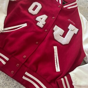 Peut inclure: Une veste de baseball rouge et blanche avec une lettre blanche "J" et le numéro "04" sur le devant. La veste a des rayures blanches sur les manches et le bas. Le nom "Lindsay" est brodé sur le côté droit de la veste.