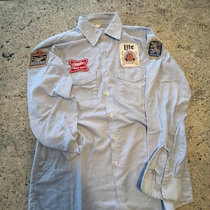 以下が含まれることがあります： ライトブルーと白のストライプのシャツで、複数のパッチが付いています。「Miller Genuine Draft」、「Miller High Life」、および「Lite」のロゴがパッチに含まれています。 シャツには2つの胸ポケットとボタン付きの袖口が付いています。