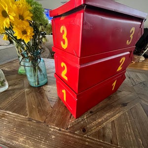 Op de afbeelding: Drie gestapelde rode metalen dozen met gele cijfers 1, 2 en 3. De dozen staan op een houten tafel met een visgraatpatroon. Een boeket gele en groene bloemen in een blauwe glazen pot staat op de achtergrond.