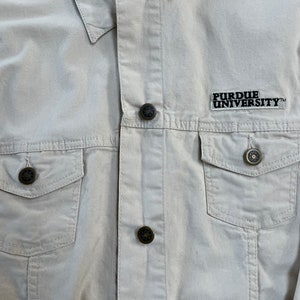 May include: A light tan jacket featuring the embroidered text "PURDUE UNIVERSITY". The jacket has two front pockets, decorative buttons, and a collar. The jacket is made of a durable fabric.