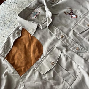 Puede incluir: Una camisa de manga larga de color gris claro con un parche de gamuza marrón en el hombro izquierdo. La camisa tiene un cuello abotonado, un bolsillo con botón único en el pecho izquierdo y dos bolsillos con solapa abotonada en la parte delantera. La camisa tiene un logotipo de cabeza de ciervo bordado en el lado derecho del pecho. El logotipo es verde, marrón y blanco. La camisa tiene el texto "RedHead" en una etiqueta verde.