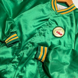 May include: Green satin baseball jacket with yellow and white striped trim. The jacket has a snap closure and a patch on the left chest that says "USA".