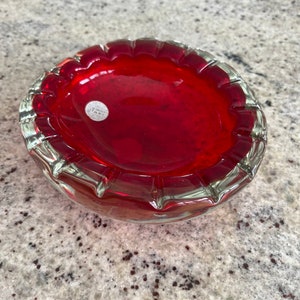 以下が含まれることがあります： 赤いガラス製の灰皿で、縁は透明でギザギザになっています。灰皿の底には「Made in Italy」と書かれたステッカーが貼られています。