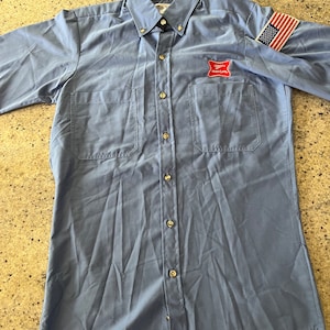 以下が含まれることがあります： ライトブルーの長袖の作業着シャツ。赤と白のロゴには「Busch Light」と表示され、右袖には小さなアメリカ国旗のパッチが付いています。