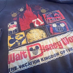 May include: A dark navy sweatshirt featuring a collage of patches with Disney themes. The design includes a red castle, Mickey Mouse, the text Walt Disney World, The Vacation Kingdom of the World, and Animal Kingdom, all stitched onto the fabric.